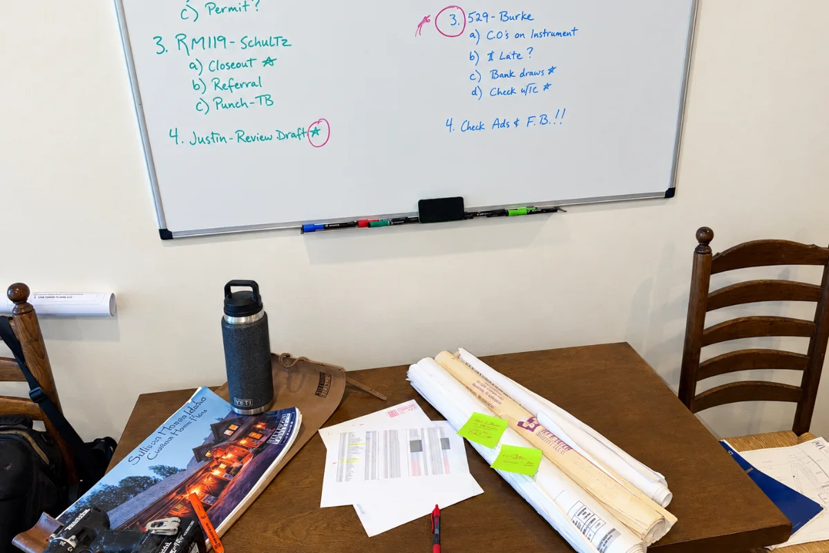 A whiteboard of jobs above a table piled with plan rolls and sticky notes, a printed spreadsheet, a plan book and a drill