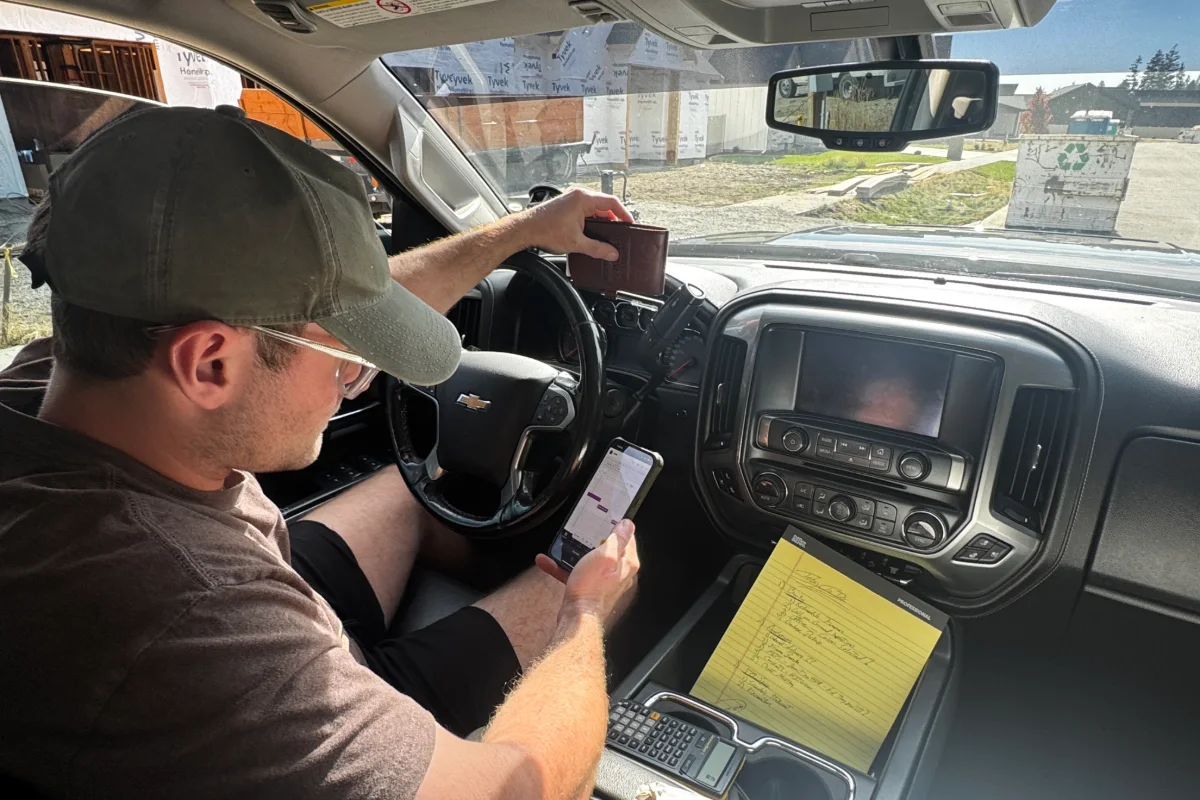 A superintendent in his pickup on a job site, checking the schedule on his phone, a yellow legal pad of notes and a calculator in the console